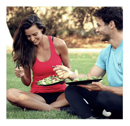 A smiling man and woman eat salads together while sitting on the grass.