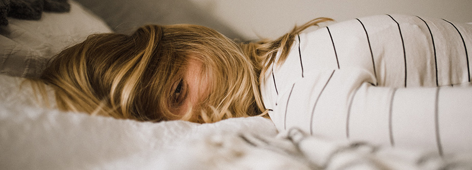 A person with blonde hair lies on a bed, their face partly covered by hair, with one eye looking out. They wear a striped shirt.